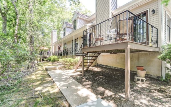 Two-story beige townhouse with a raised deck, black iron railing, and outdoor seating facing a tree-filled yard.