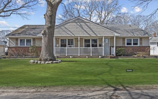Front view of a ranch-style house with a wide porch, white railings, brick base, and a large leafless tree in the yard.