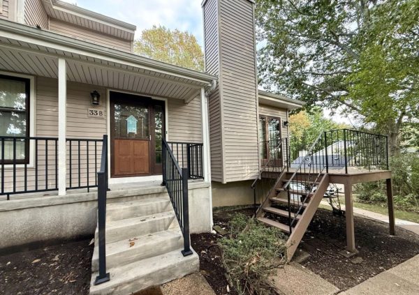 Front entrance of a beige apartment building with a brown door, concrete steps, and black metal railings.