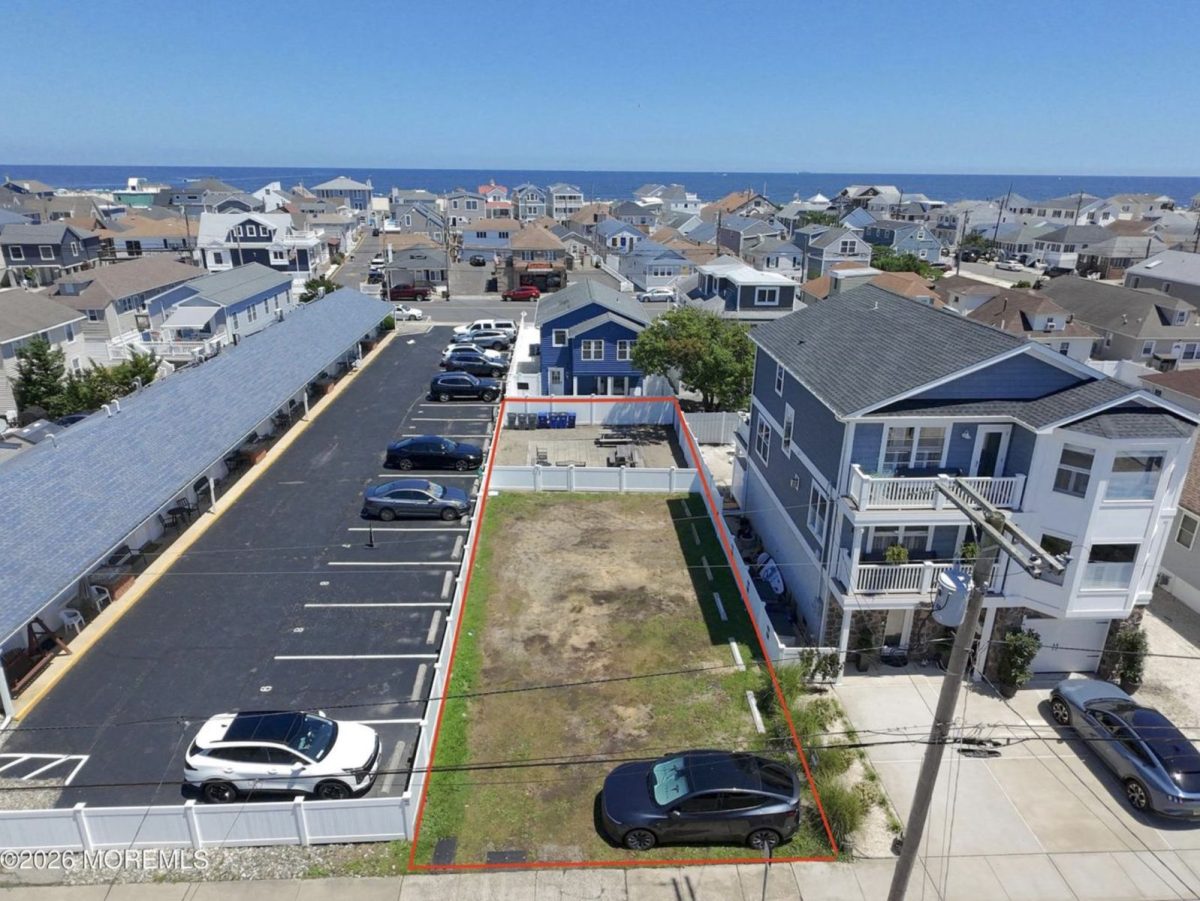 Aerial view of a coastal neighborhood with a rectangular vacant lot outlined in red between a blue house and a parking area, ocean in the distance.
