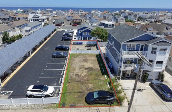 Aerial view of a coastal neighborhood showing a rectangular vacant lot outlined in red, between a blue-gray house and a parking lot, with the ocean in the distance.