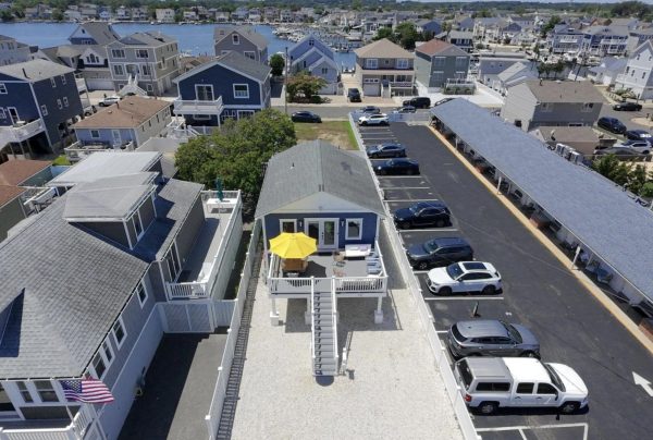 Aerial view of a coastal neighborhood with multi-story beach homes, a central blue house with a yellow patio umbrella, and a long parking lot nearby.